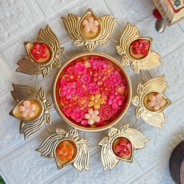 DH-01: Seven lotus diyas on a gold metal stand, around an urli of pink and red blooms, on a white table.